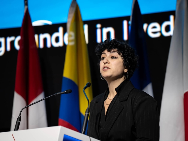 A woman standing at a lectern speaking, there are flags and a large screen behind her