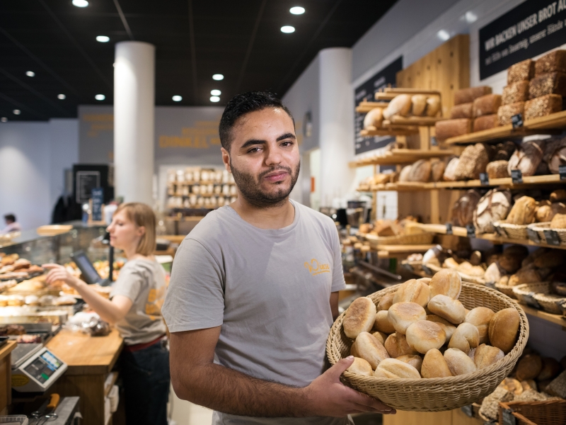 A baker stands with showing a basket of bread behind a shop counter