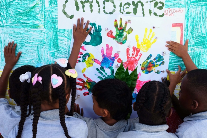 Children painting a picture with their hands. The photo is taken from behind the 5-6 children, and they have made handprints in different colours on a piece of paper, with the word 'ambiente' painted in what looks like fingerprints.