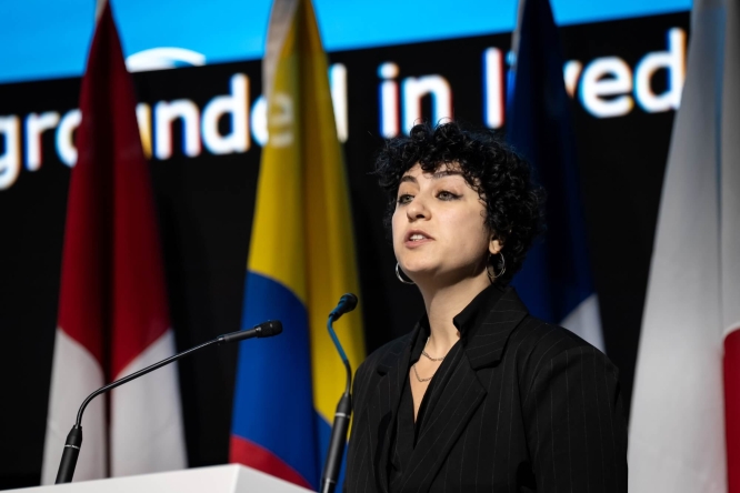 A woman standing at a lectern speaking, there are flags and a large screen behind her