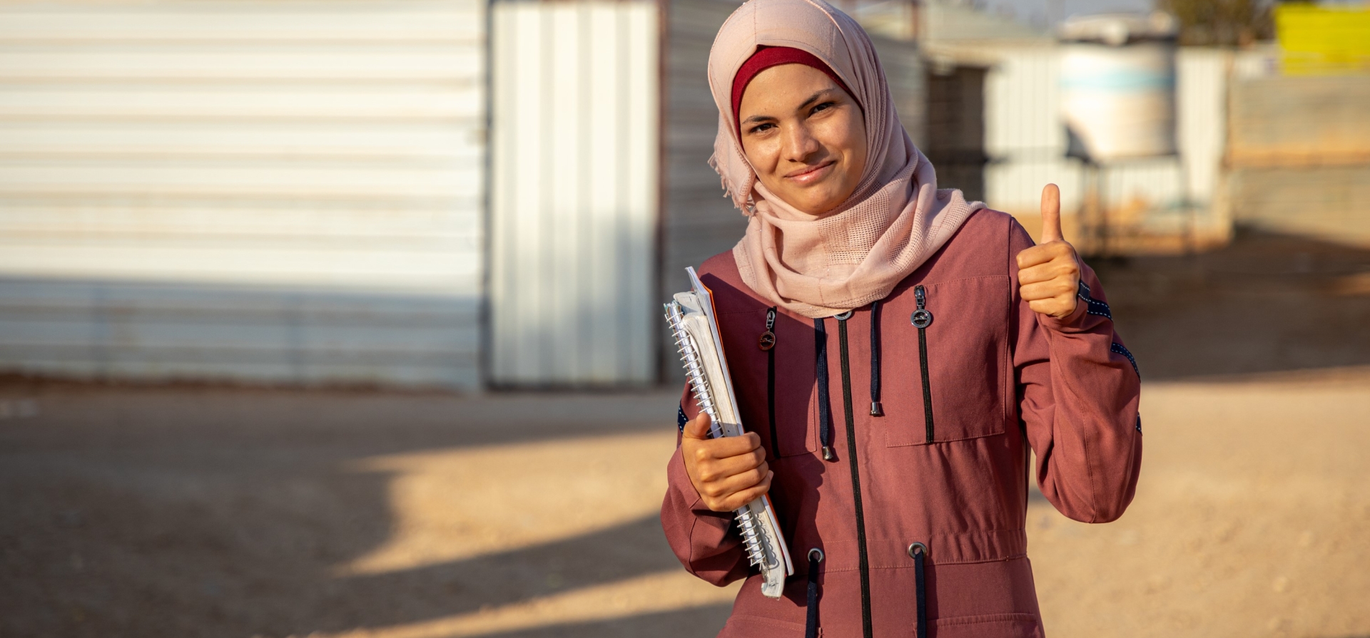 A woman standing outside, with buildings behind her, smiling and giving a thumbs up to the camera. She is holding textbooks and notebooks under one arm