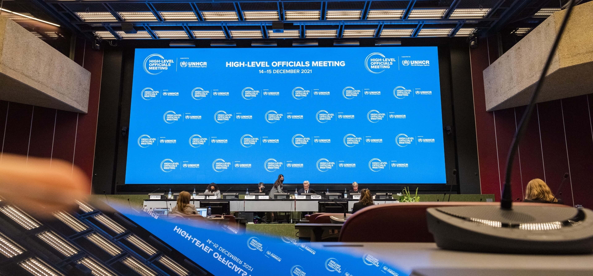 A view from the back of the room towards the stage, where there is a large screen with the High-Level Officials Meeting logo repeated across it. This, and the ceiling, are reflected in the foreground of the image, where a laptop or something is sitting