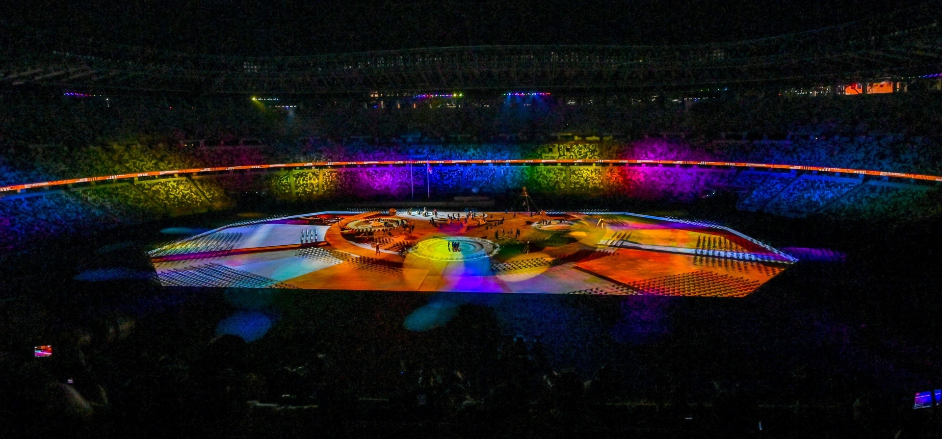 Inside the stadium, it is dark, except for the centre which is lit up with a lot of different coloured bright lights. In the middle, there are people on the stage, and the stands are filled with crowds