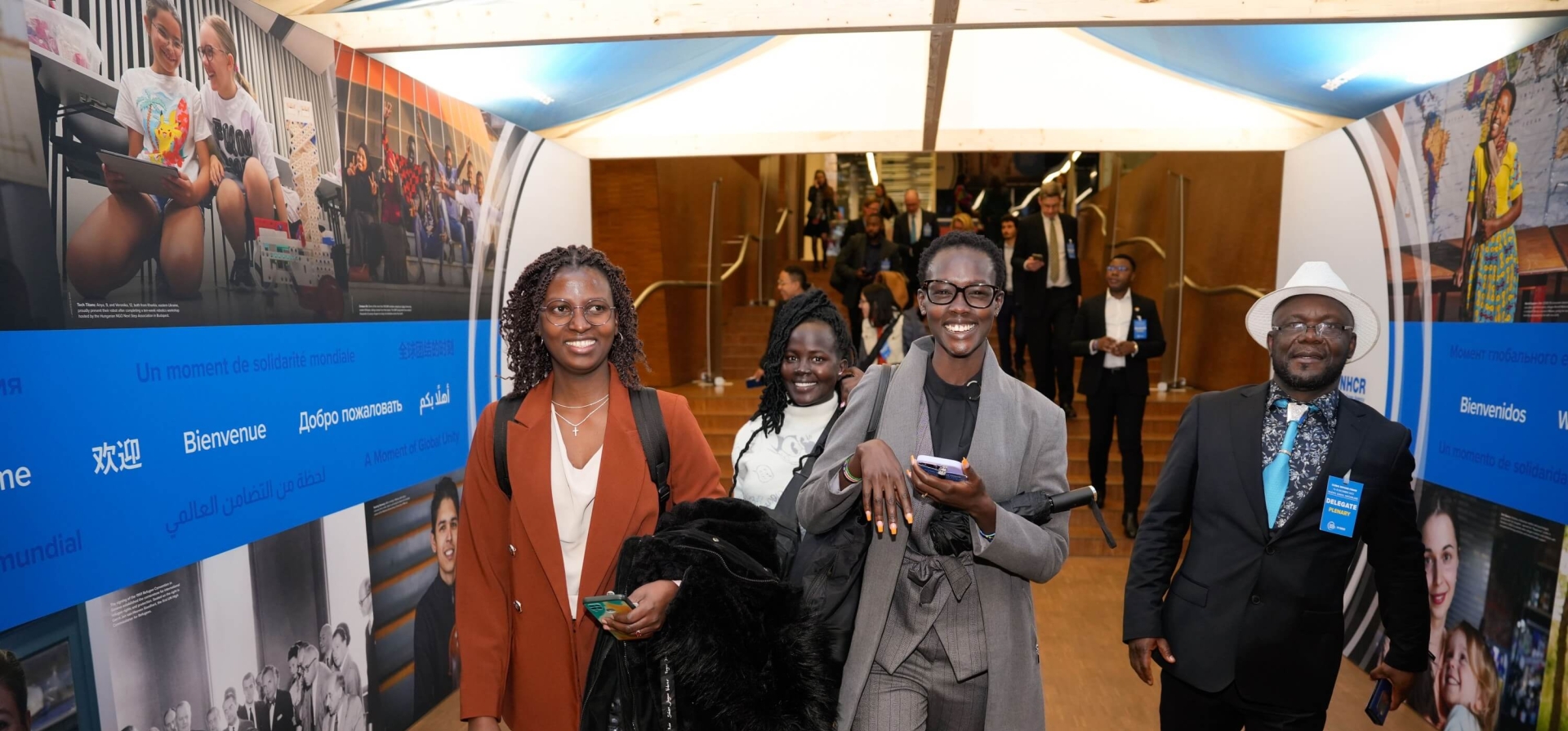 Four people walking through the entry tunnel - an indoor structure within the conference centre with images and the word "Welcome" in different languages on the wall and a tented roof. The four people are smiling at the camera. Behind them more people are descending a staircase coming towards the walk way
