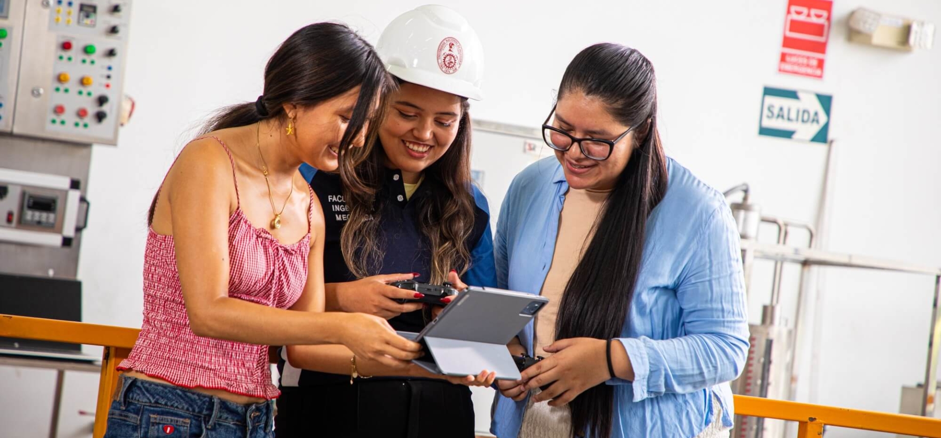 Three women standing around a tablet, which one of them is holding. The woman in the middle is also holding a remote control (similar to those used for remote control cars) and wearing a hard hat.