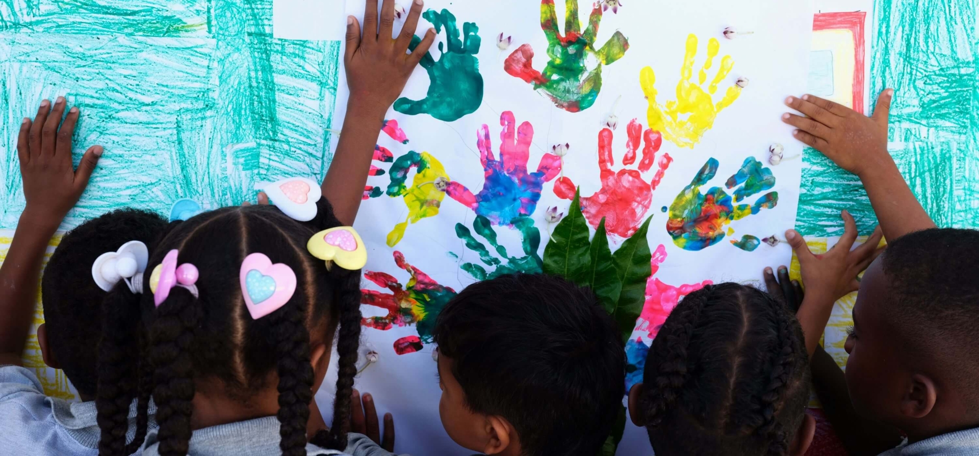 Children painting a picture with their hands. The photo is taken from behind the 5-6 children, and they have made handprints in different colours on a piece of paper, with the word 'ambiente' painted in what looks like fingerprints.