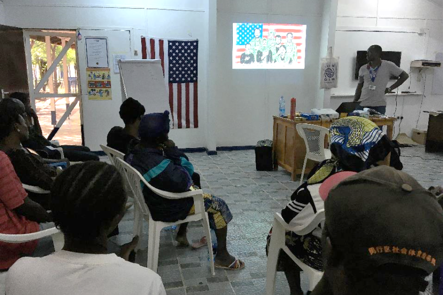People seating on chairs in a room, listening to someone giving a presentation.