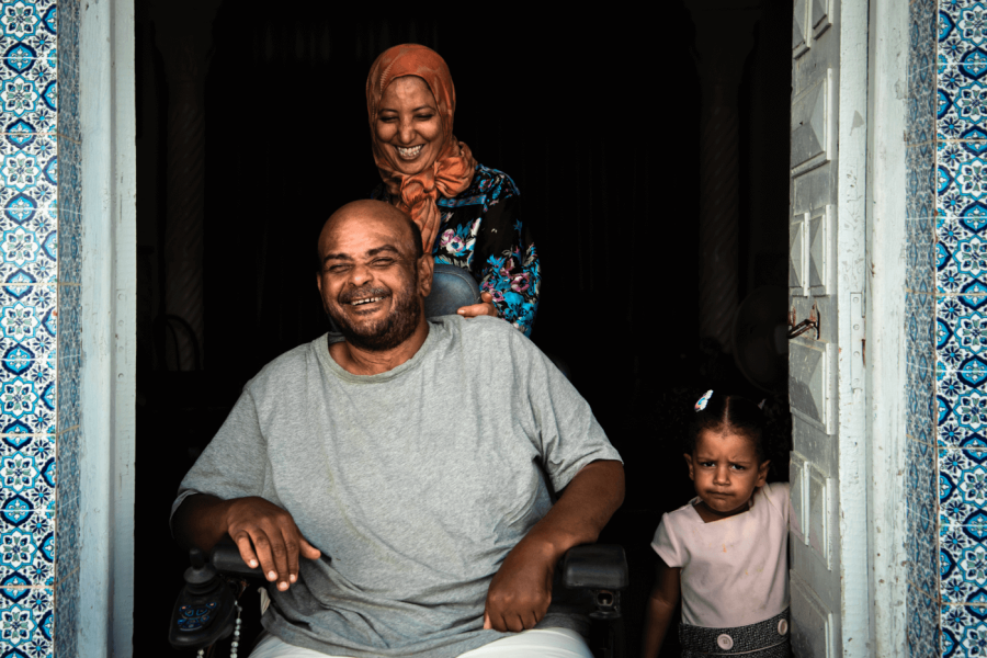 Family in a doorway, man is in a wheelchair with woman standing behind him. They are both laughing. Next to them, a little girl frowns at the camera