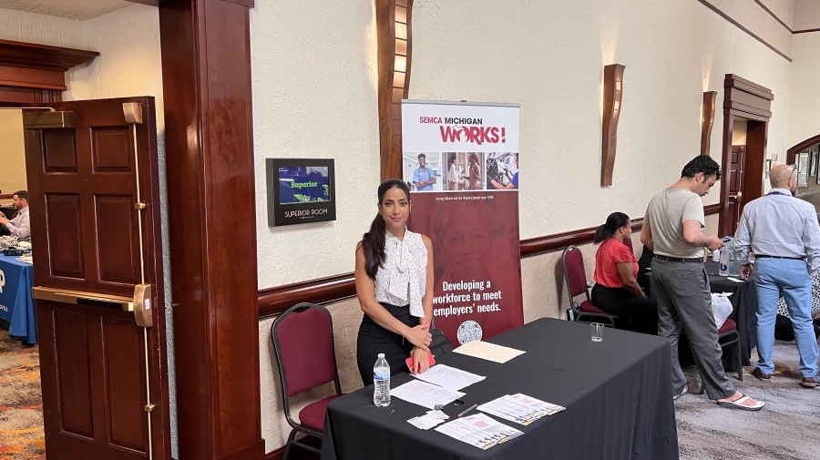 A woman stands and smiles at the camera. In the background there is a roll-up banner for SEMCA, she has in front of her a table with forms.