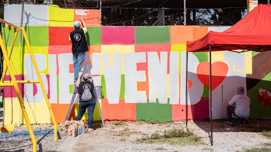 Three people are painting a mural that reads "Bienvenidos"