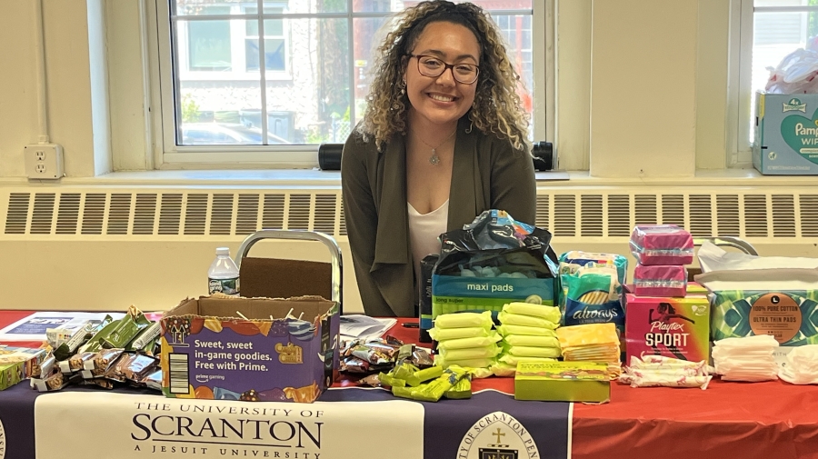 A woman sits at a booth with a Scranton logo. There are menstrual products on the table.