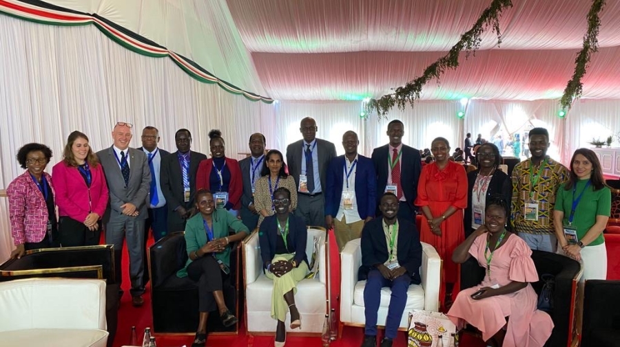 A group photo of refugee participants and UNHCR staff. Some are sitting in armchairs, others standing behind, all smiling and looking at the camera