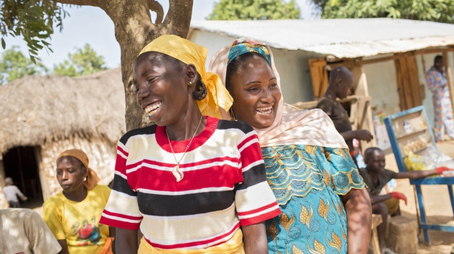 Two women standing together under a tree laughing. There are other people and buildings in the background