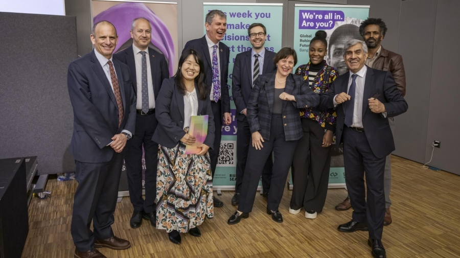 A group photo of nine people smiling and laughing - two of them are putting their elbows together. They are standing in front of banners advertising events at the Global Refugee Forum