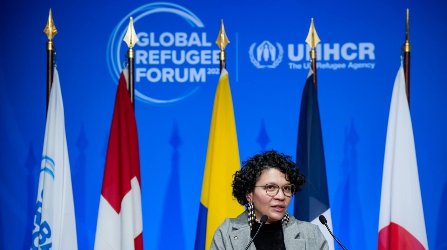 A woman speaking at a podium with eight different flags behind her and the Global Refugee Forum logo on the wall behind the flags