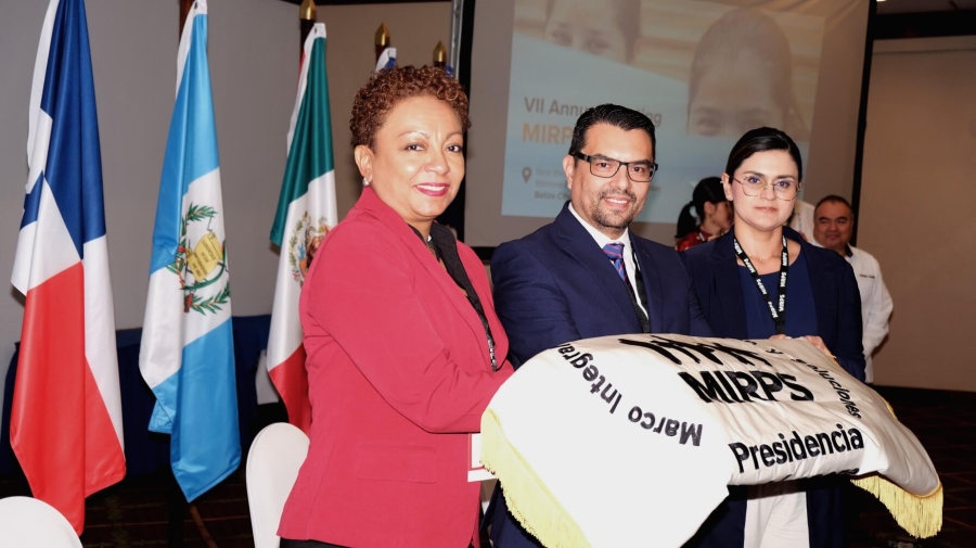 Three people standing together with a folded piece of cloth showing the MIRPS logo with "Presidencia" written on it - behind them are some of the flags of the MIRPS States