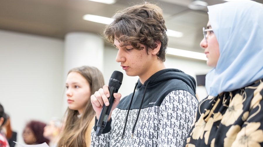 Three youths sitting in a row with other people listening behind them. The one in the middle is speaking into a microphone
