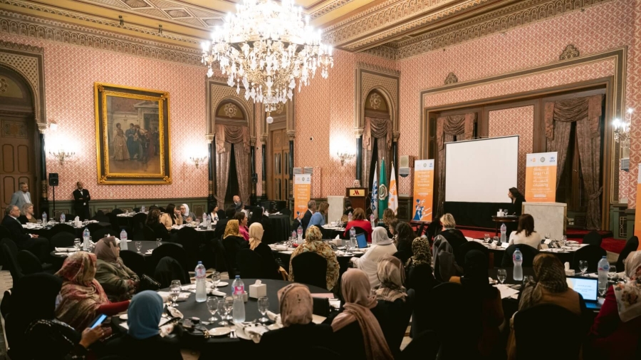 A meeting in what looks like an event room of a grand hotel - there are six round tables around which people are sitting, a stage with a Podium where someone is speaking, and a chandelier coming from the ceiling.