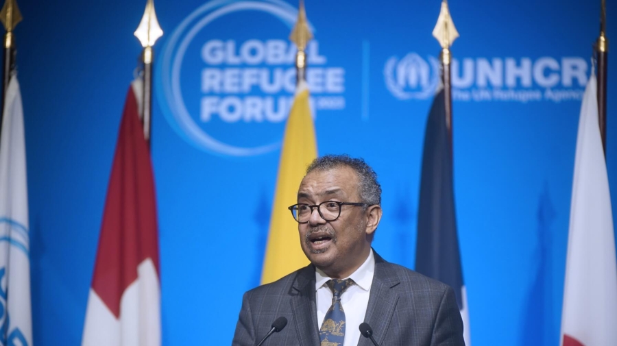 A man standing at a lectern speaking, there are flags behind him with the Global Refugee Forum logo on the wall behind
