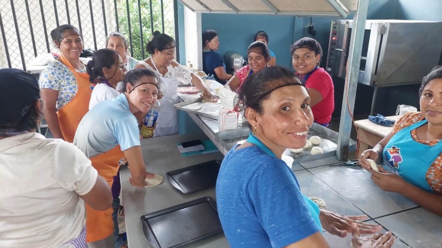 A group of twelve women standing and sitting around a long work table kneading dough.  A couple of them are looking towards the camera, smiling