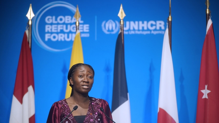 A woman standing at a podium speaking into microphones. Behind her are five flags, and behind the flags a blue background with the Global Refugee Forum logo on it