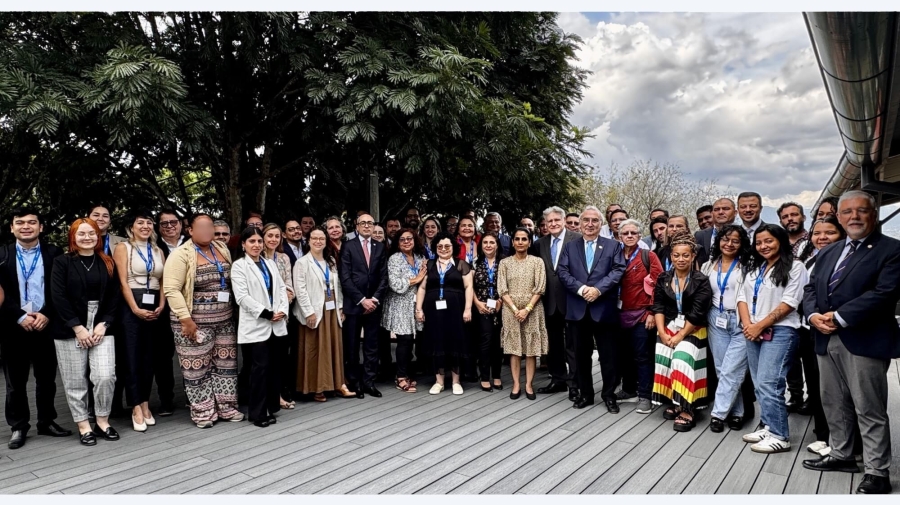 A group photo of around 50 people standing on some wooden decking. There is a tree directly behind those to the left, and behind those on the right is a cloudy, but bright sky.