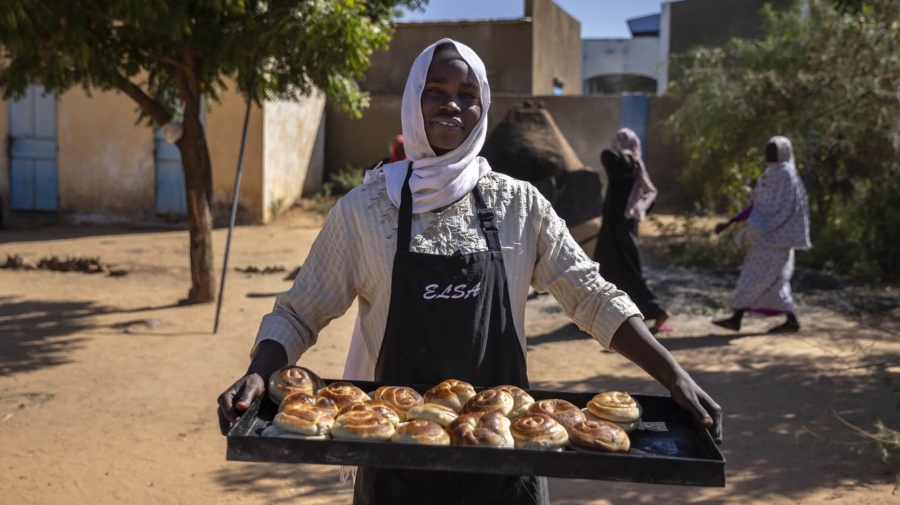 A woman standing holding a tray of baked swirls (cinnamon or raisin type of pastry) - she is wearing an apron, smiling at the camera, standing outside with a tree and some buildings in the background as two people walk behind her