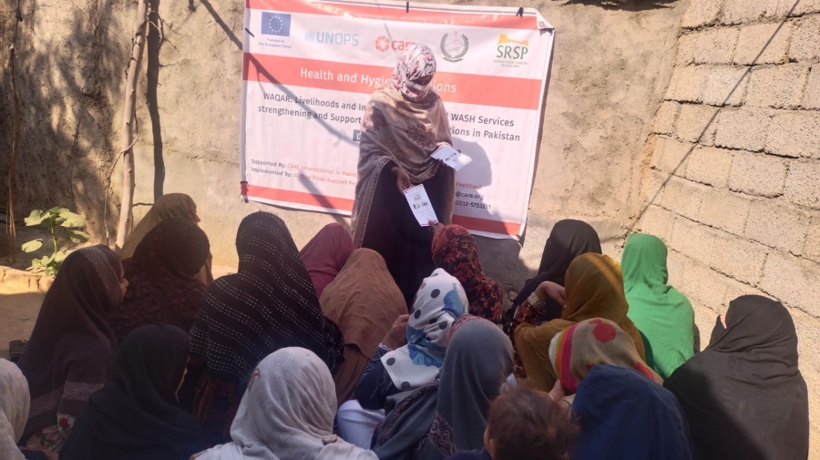 A group of women sitting with one standing at the front handing out leaflets. Behind the one at the front is a banner saying "Health and Hygiene" - they are outside with sand-coloured walls around them
