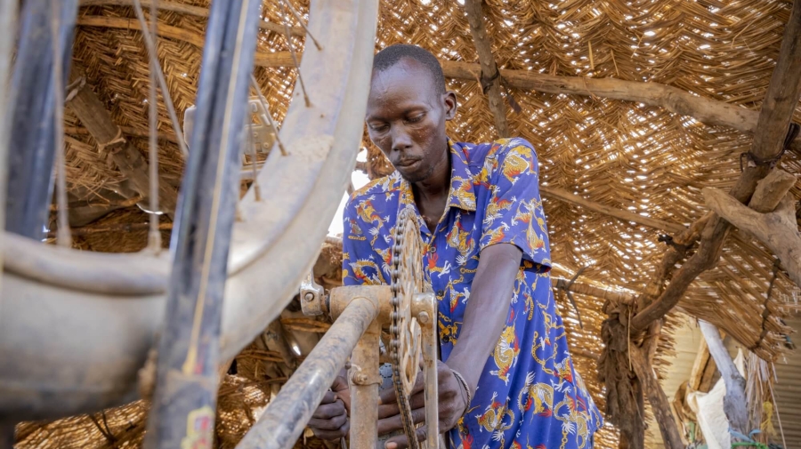 A man is repairing a bike, which is turned upside down. In the foreground is one of the bikes wheels, with the pedals and cogs visible behind, further back, the man is leaning over the back wheel of the bike, holding it with one hand and what looks to be a screwdriver in the other. Overhead there is a thatched roof held up with large logs