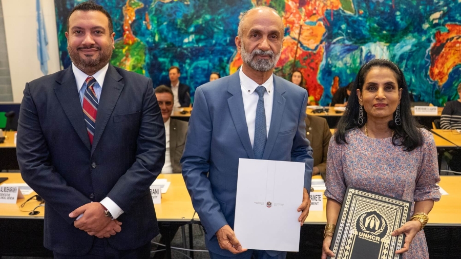 Three people standing next to one another, with a bright abstract painting on the wall behind them. The man in the centre is holding a document with a coat of arms on it. The woman on the right is holding a black and gold plaque with UNHCR's logo on it