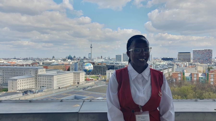 Rebecca standing on the balcony of German Federal Ministry for Economic Cooperation and Development, behind her is the Berlin skyline, with the TV Tower (Fernsehturm) and Berlin Cathedral (Berliner Dom) in the distance and the Welt hot air balloon in the foreground. The sky is blue with white/light grey clouds.