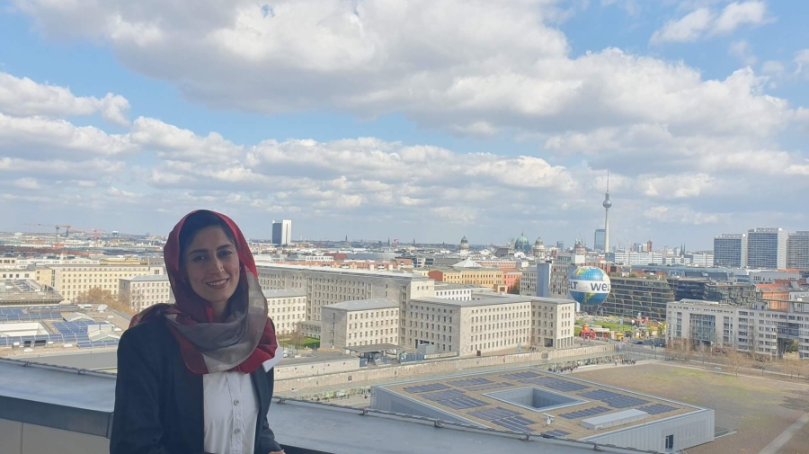Safia standing on the balcony of German Federal Ministry for Economic Cooperation and Development, behind her is the Berlin skyline, with the TV Tower (Fernsehturm) and Berlin Cathedral (Berliner Dom) in the distance and the Welt hot air balloon in the foreground. The sky is blue with white/light grey clouds.