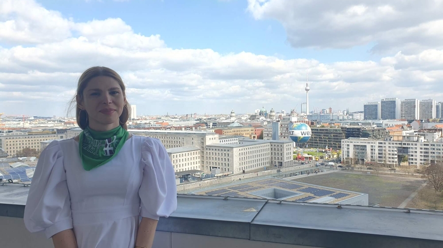 Vlada standing on the balcony of German Federal Ministry for Economic Cooperation and Development, behind her is the Berlin skyline, with the TV Tower (Fernsehturm) and Berlin Cathedral (Berliner Dom) in the distance and the Welt hot air balloon in the foreground. The sky is blue with white/light grey clouds.