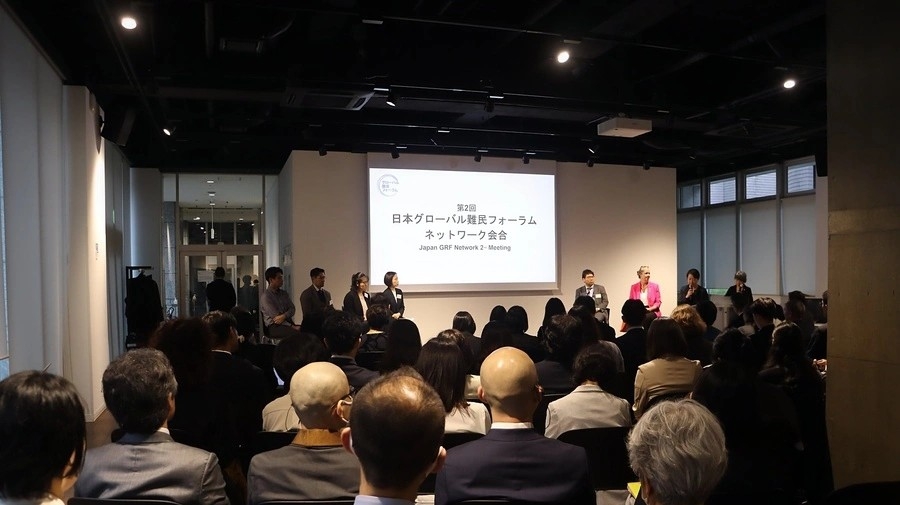 A photo from the meeting, taken at the back of the room, showing participants sitting down, with eight people standing at the front facing them, the lights shining on them, and a screen behind them saying with some writing in Japanese, and in English the words "Japan GRF Network 2nd Meeting"
