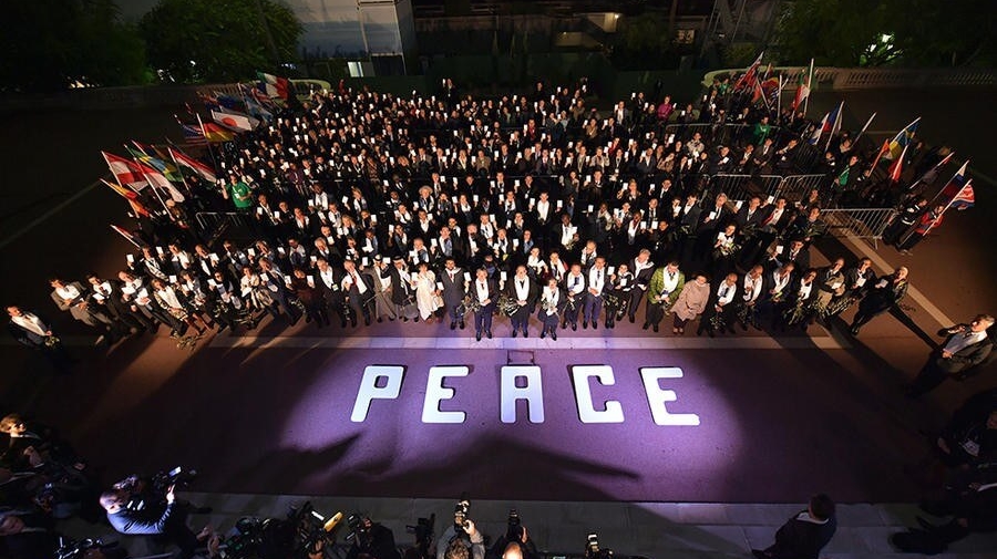 An aerial view of a large group of people standing outside, with a purple carpet and word peace written on the ground in front of them. There are flags near the people, and on the other side of the carpet are photographers and videographers