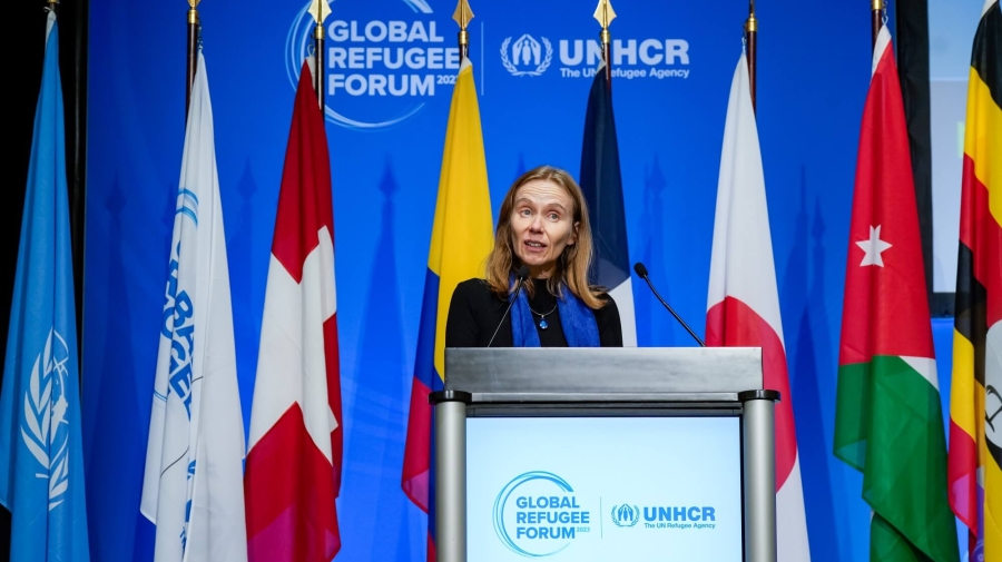 A woman standing at a podium with the Global Refugee Forum logo on it. Behind her are flags on poles, and behind those a blue backdrop with the Global Refugee Forum logo on it.