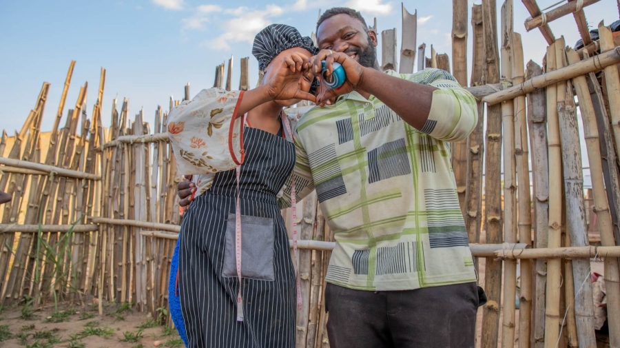 A man and a woman standing together outside, with a wooden fence behind them. They have their arms around one another, and are making a heart shape with his left hand and her right.