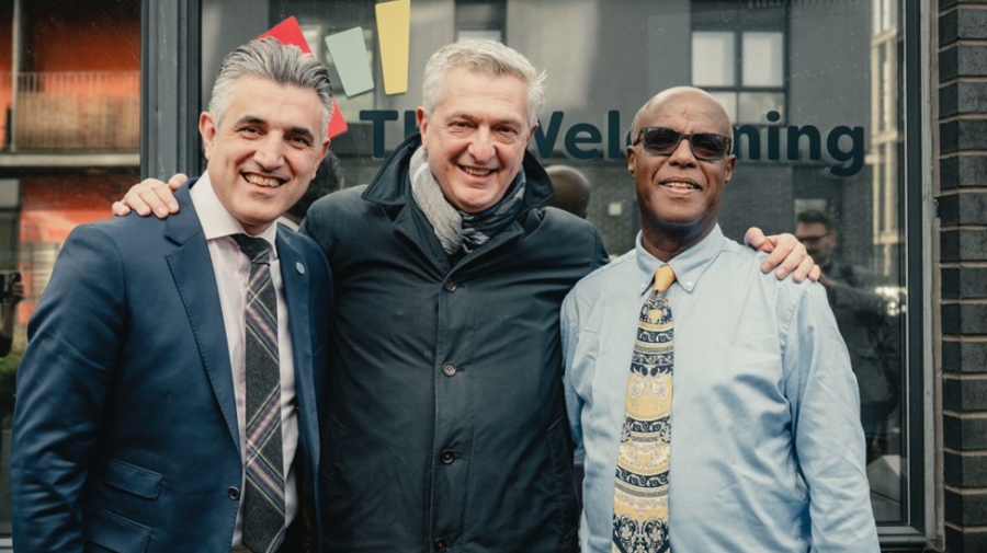 Three men standing together outside on what looks like a street in the centre of a town. The man in the middle has his arms around the shoulders of the other two. They are all smiling