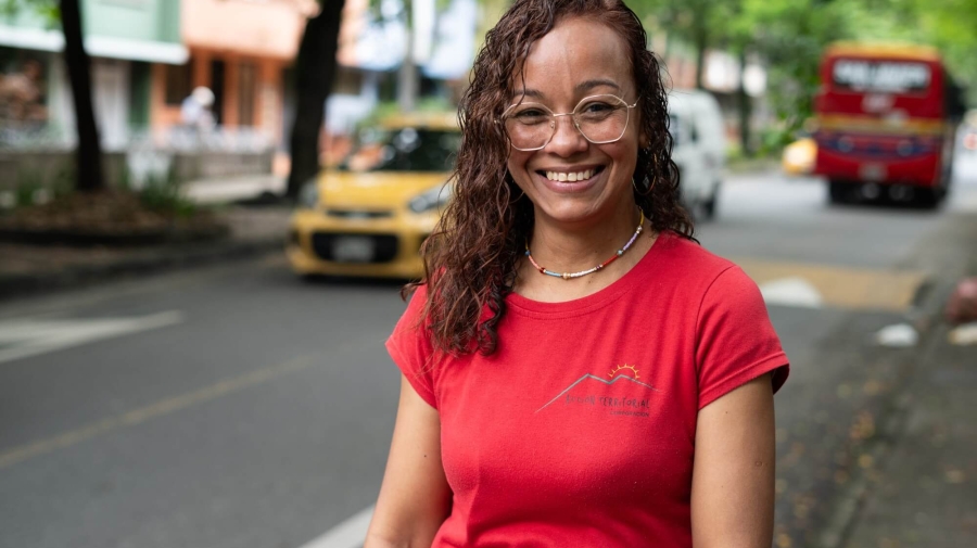 A woman standing in a tree-lined street, smiling at the camera. Behind her, there is a car driving one way and a bus driving in the other direction