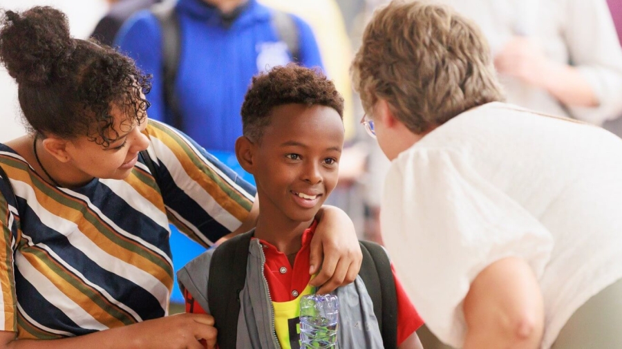 A boy being greeted by a woman, who is bending down to speak to him. He is holding a bottle of water upside down, smiling, while his mother has her arm around his shoulders and is looking at him