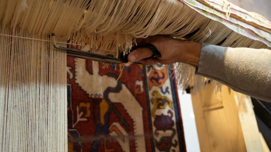 A rug being woven - it is hanging down, and a hand is holding scissors trimming the threads