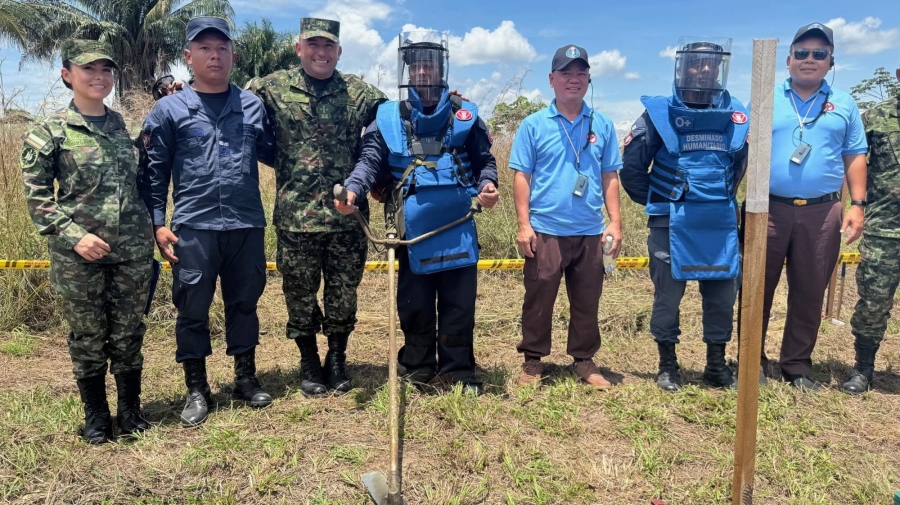 Eight people standing side by side in a field. They are dressed in a mixture of army fatigues and bomb-disposal type safety clothing, including helmets. They have tools, that look a bit like rakes, on the ground in front of them. The sky behind them is blue.