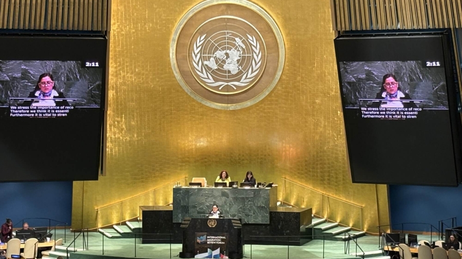 The Mexican Minister delivering the statement at the UN headquarters in New York. She is standing on a stage, with a large golden wall behind her with the UN logo on it. To the sides of the wall are large screens showing her speaking.