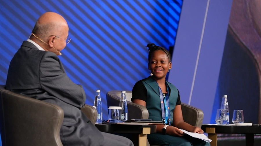 The High Commissioner and Ms. Magesa sitting in chairs on stage. The photo is taken from the side, showing the side and back of the High Commissioner, and the face of Ms. Magesa. She is smiling.