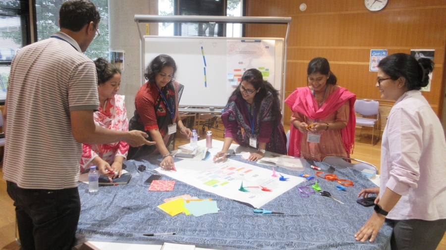 A group of six people standing round a table putting colourful pieces of paper (perhaps some origami?) on a large sheet of paper