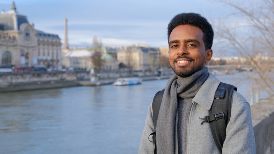 A man standing next to a river. He is smiling at the camera. On the other side of the river are some grand buildings, and in the distance is a boat.