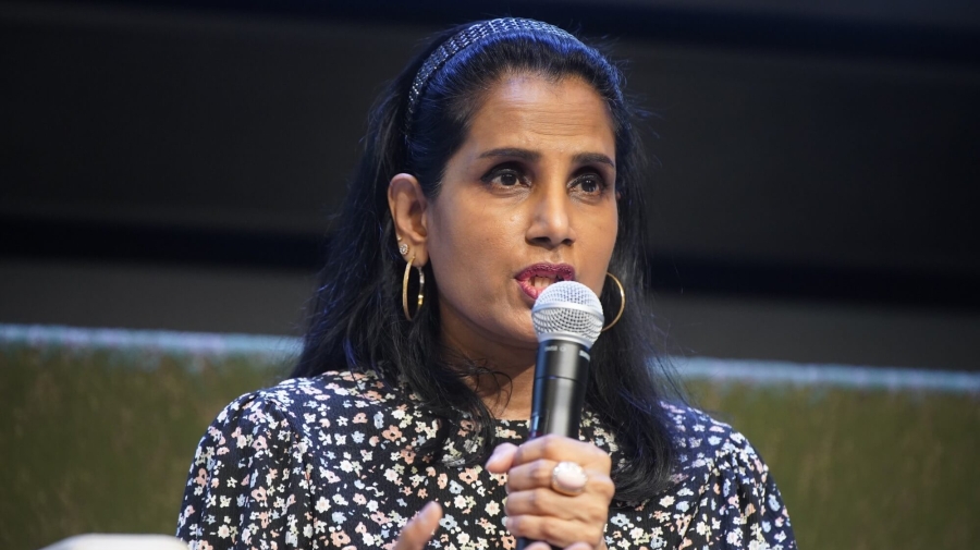 Ruven speaking into a microphone. A close up image only showing her upper body and torso, she is wearing a floral shirt, and holding out one hand to gesture while speaking.