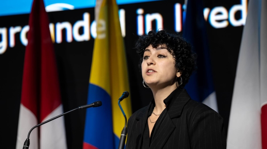 A woman standing at a lectern speaking, there are flags and a large screen behind her