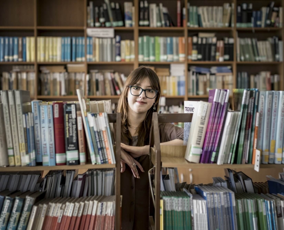 Olha standing in a library, leaning between two bookcases, looking at the camera